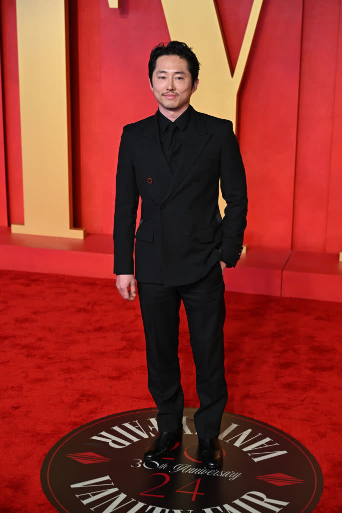 A man stands on a red carpet at an event, wearing a formal black suit with a boutonniere