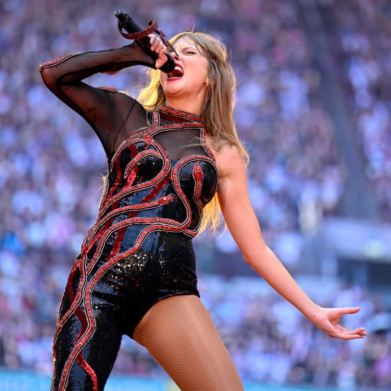 Taylor Swift performs passionately on stage, wearing a sparkling, asymmetrical black outfit with red accents and sheer detailing