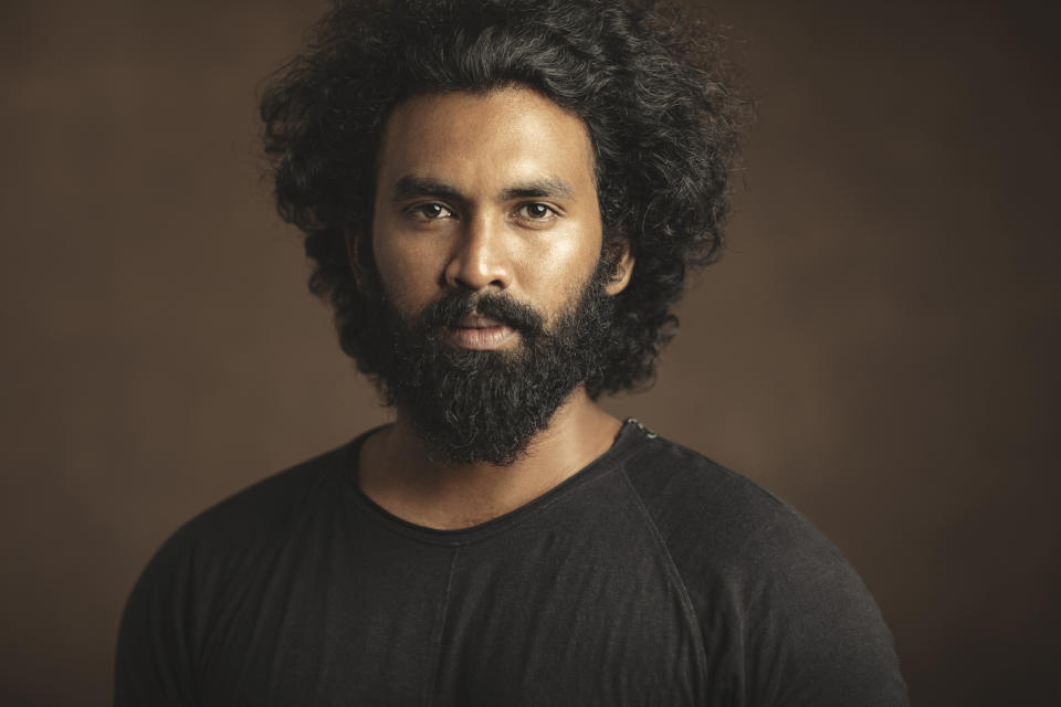 A man with a full beard and curly hair looks directly at the camera, wearing a plain black T-shirt against a neutral background. No other persons are in the image
