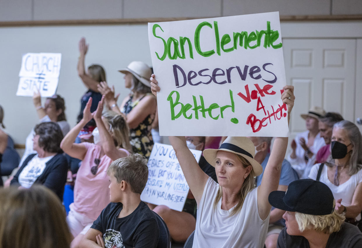 A woman in a straw bowler holds a sign above her head saying: San Clemente Deserves Better, Vote 4 Betteh, with other signs visible saying: Church does not equal State..