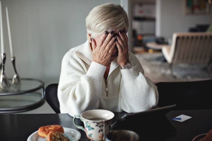 An older woman covers her face with her hands, showing emotion and resting her elbows on a table, with a tablet and breakfast on it