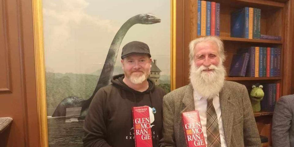 Loch Ness enthusiast Alan McKenna (L) with noted Loch Ness researcher Adrian Shine (R). Both hold bottles of whisky and stand in front of a painting of the Loch Ness Monster