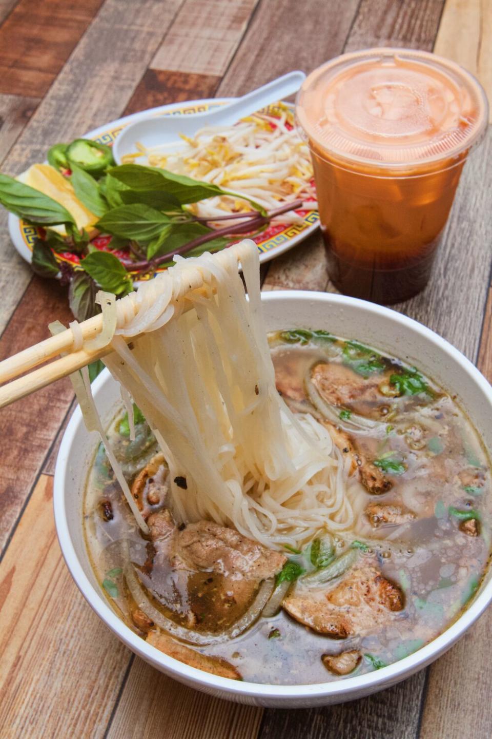 Les baguettes soulèvent les nouilles de riz du pho au porc carbonisé. Derrière se trouve une tasse de thé glacé thaïlandais et une assiette de basilic et de germes de soja.