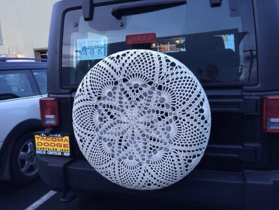 A Jeep with a crocheted cover on the spare tire is parked in a lot. A yellow Tacoma Dodge dealership plate is attached to the spare tire cover