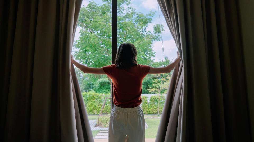 Person with short hair stands inside and holds open large curtains, facing a window looking out at greenery