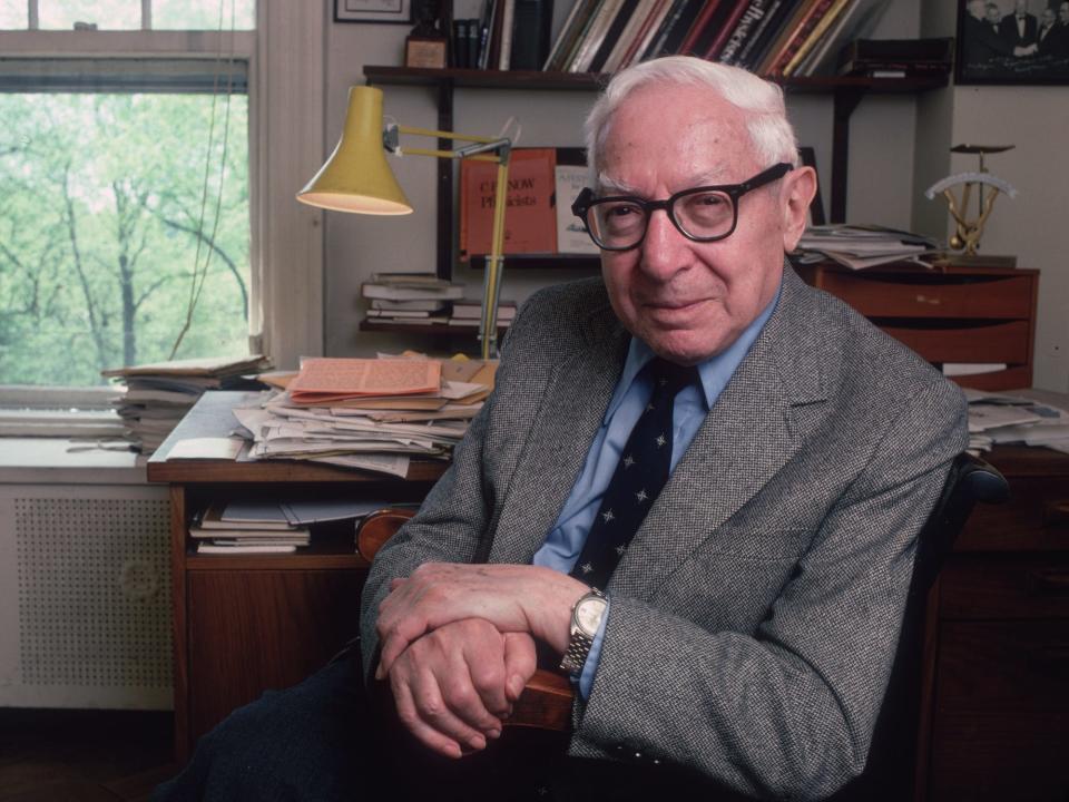 Nobel Prize winner Isidor Isaac Rabi sits in front of a desk and a window wearing black framed eyeglasses and a tweed jacket.