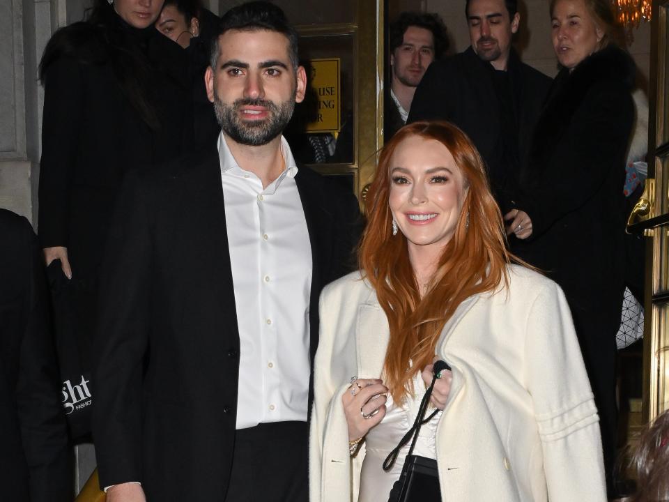 bader shammas and lindsay lohan posing together on red steps outside of a hotel. lohan is in a white gown and jacket, smiling, and shammas is wearing a white shirt and black suit