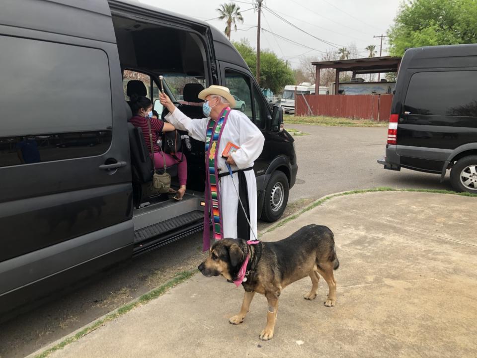 A masked priest in robes and cowboy hat accompanied by a dog on a leash gives blessings near the open door of a van