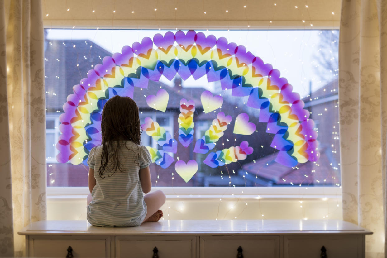 A little girl is looking out of a window at home.  She is sitting under a rainbow and fairy lights which have been displayed on the glass.  Stay at home social media campaign for coronavirus prevention.