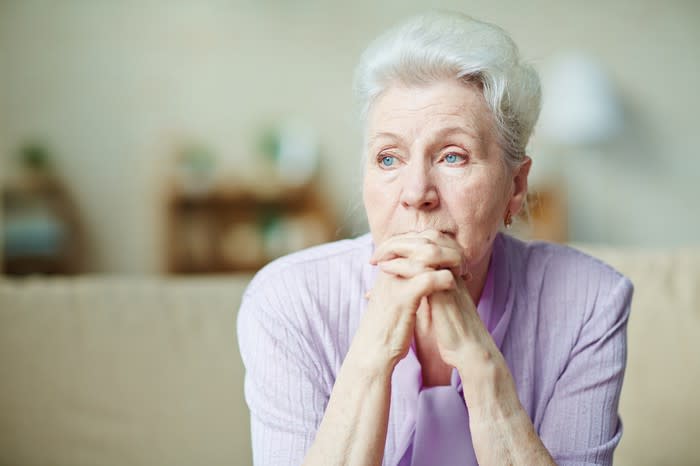 Older woman with clasped hands sitting on a couch and looking worried.