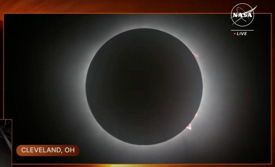 screengrab from NASA live shows total solar eclipse dark disc of the moon with a few pink prominences around the edge in the glow of sunlight