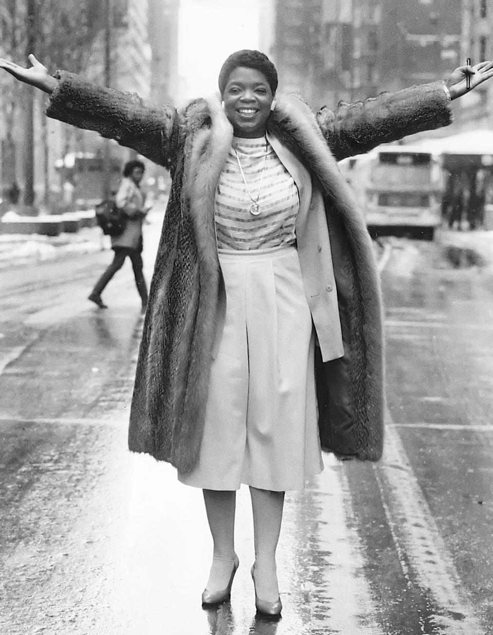 Oprah Winfrey stands in the middle of a city street with arms outstretched, wearing a fur coat and smiling. Snow is on the ground, and buildings are in the background