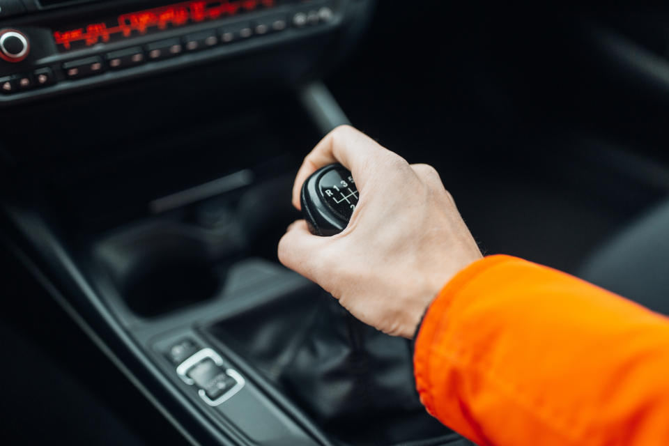 A hand wearing an orange sleeve is gripping the gear shift of a manual transmission car. The car's interior is partially visible