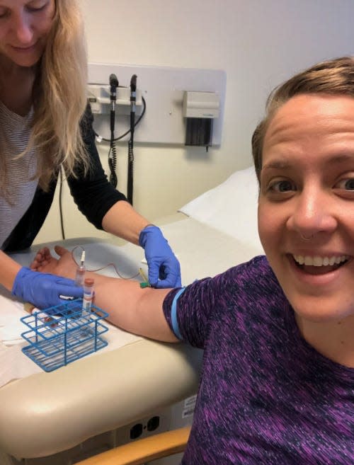 A woman in a purple shirt smiles as another woman draws blood from her arm.