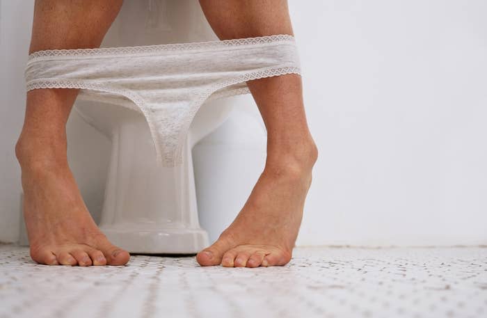 Person sitting on a toilet with underwear down to ankles, feet visible on a tiled bathroom floor