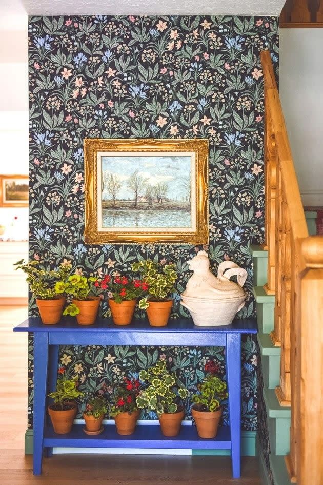 a painting on a wallpapered wall and a blue console with potted plants under a staircase