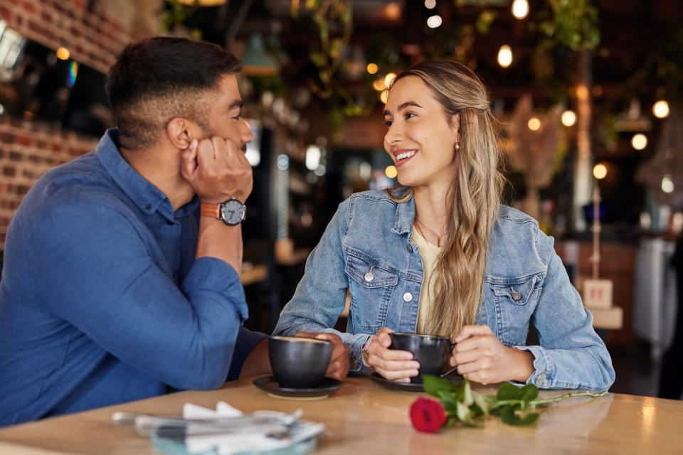 A couple in denim jackets hold coffee cups and smile at each other at a cozy cafe table with a single red rose on it