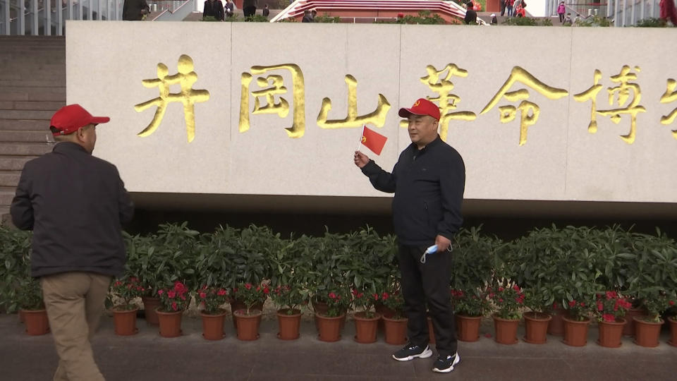 A tourists holds a flag as he poses for photographs in front of the sign for the Jinggangshan Revolution Museum in Jinggangshan in southeastern China's Jiangxi province, on April 8, 2021. On the hundredth anniversary of the Chinese Communist Party, tourists in China are flocking to historic sites and making pilgrimages to party landmarks. (AP Photo/Emily Wang)