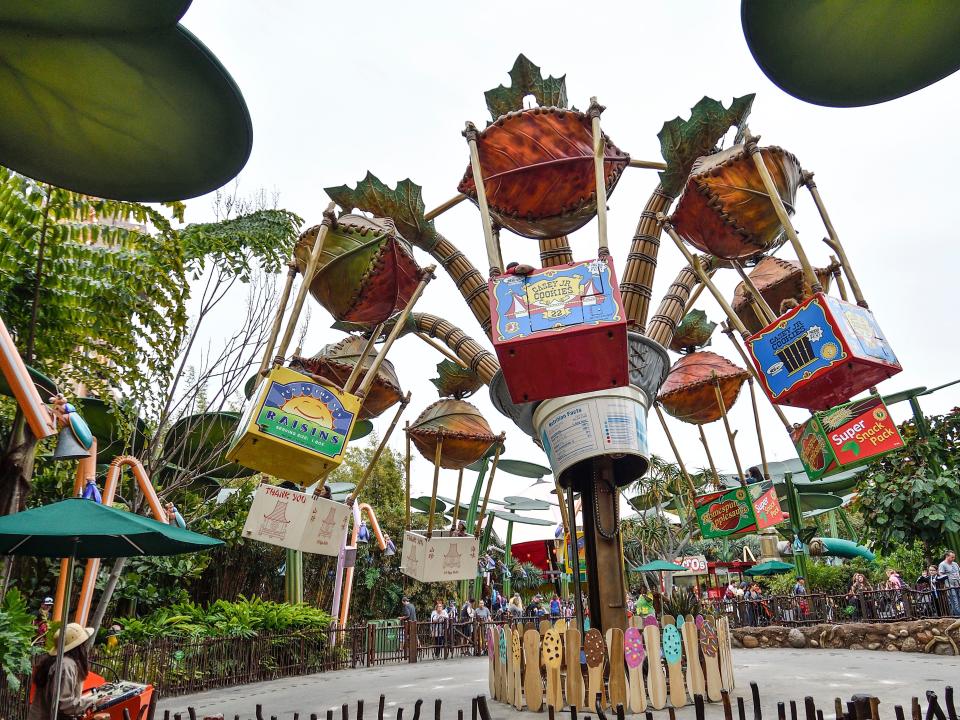 A ride with buckets holding passengers spinning in the air around a center structure. Oversized leaves and greenery surround it.