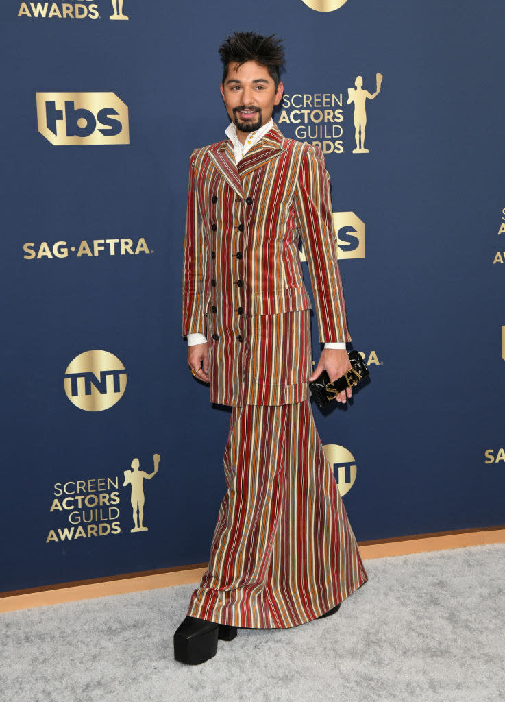 mark wears a long, warm-toned peacoat and a full-length skirt and a matching stripped pattern, and he has platform heels and a clutch that says "sex" in metallic letters