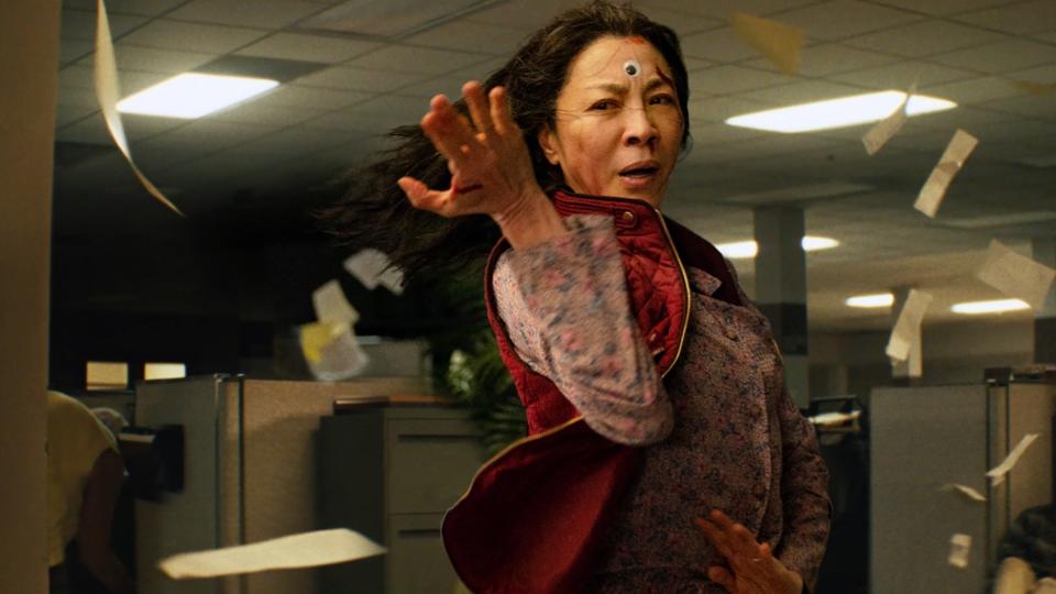 Michelle Yeoh dramatically posing in an office setting with papers flying around her in a scene