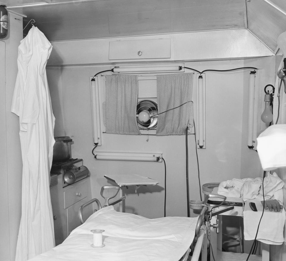 The interior of a trailer showing a hospital bed with stirrups and a white surgical gown hung from a hook.