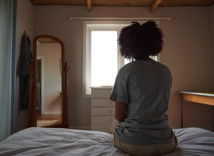 A person with curly hair sits on a bed facing away from the camera in a dimly lit bedroom with a window, mirror, and dresser in the background