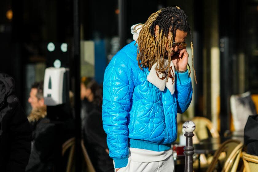 PARIS, FRANCE - JANUARY 20: A guest wears black sunglasses, a beige fluffy with brown large LV logo print pattern scarf, a blue embossed LV monogram print pattern puffer jacket, beige sport pants, outside Louis Vuitton, during Paris Fashion Week - Menswear F/W 2022-2023, on January 20, 2022 in Paris, France. (Photo by Edward Berthelot/Getty Images)