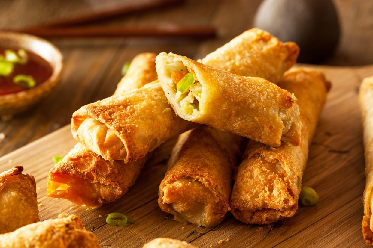 Five chinese egg rolls in a pile on a wooden cutting board with more rolls and a blurred background of chopsticks and dipping sauce