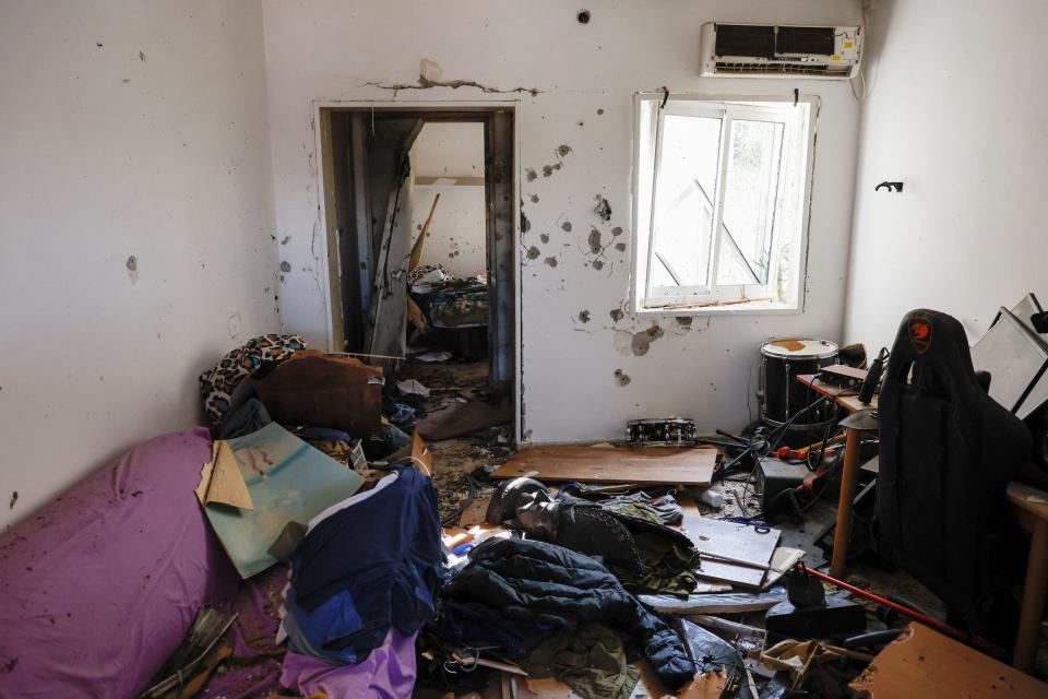 An image of a room with furniture, instruments, and clothes strewn on the floor and bullet holes all over the wall.
