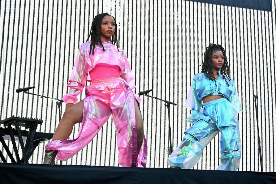 chloe bailey and halle bailey of chloe x halle pose on a stage during a music concert, chloe wears a iridescent pink outfit and halle wears a similar iridescent blue outfit, behind them is a giant screen with black and white stripes as well as microphone stands and music equipment