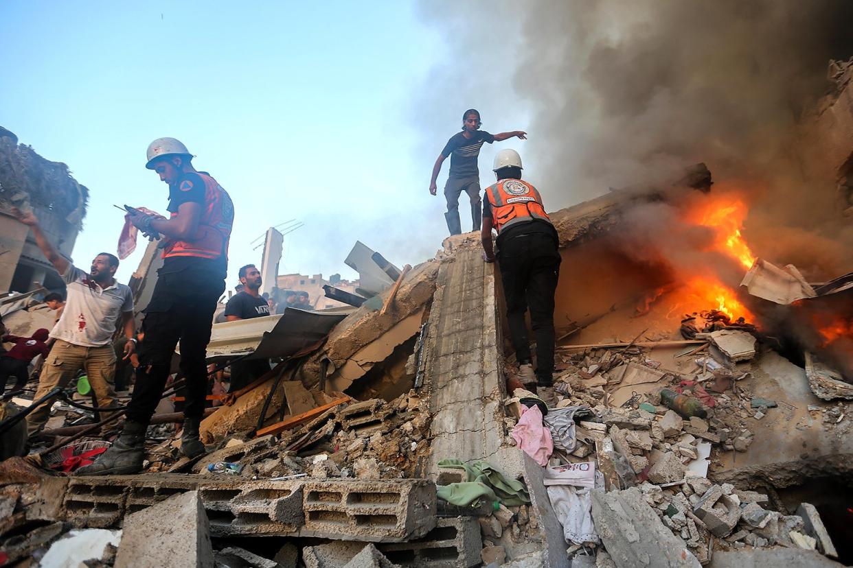 People wearing helmets and orange reflective vests climb over rubble that is partially on fire.