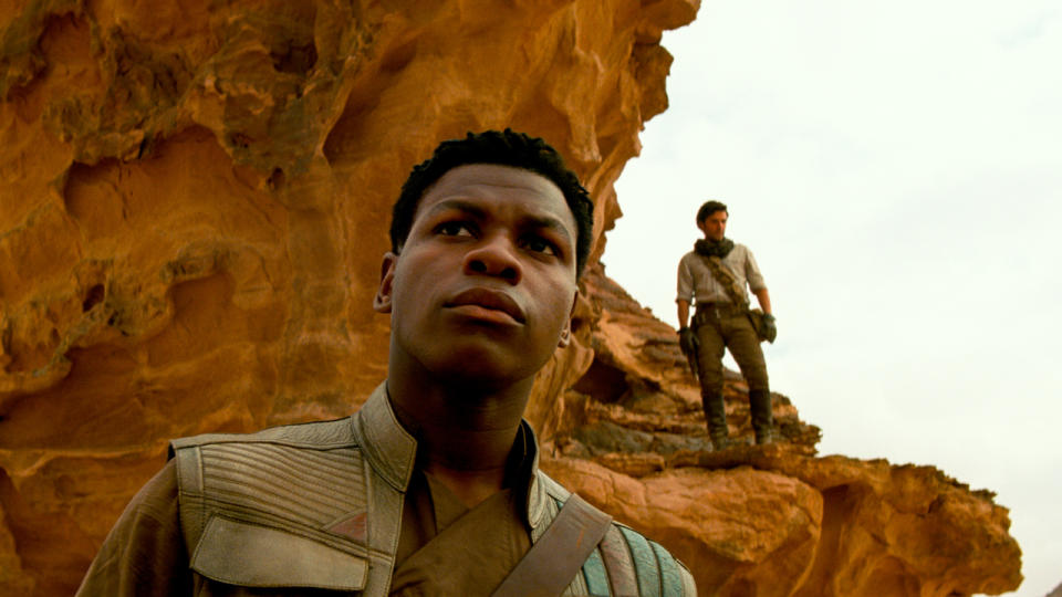 John Boyega standing near a rock formation with Oscar Isaac in the background in Star Wars: The Rise of Skywalker.