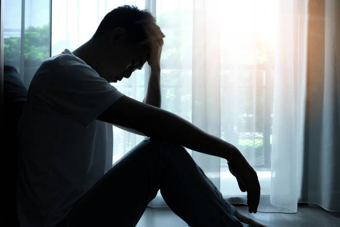 A person sits on the floor by a window, holding their head in one hand, appearing to be in deep thought or distress