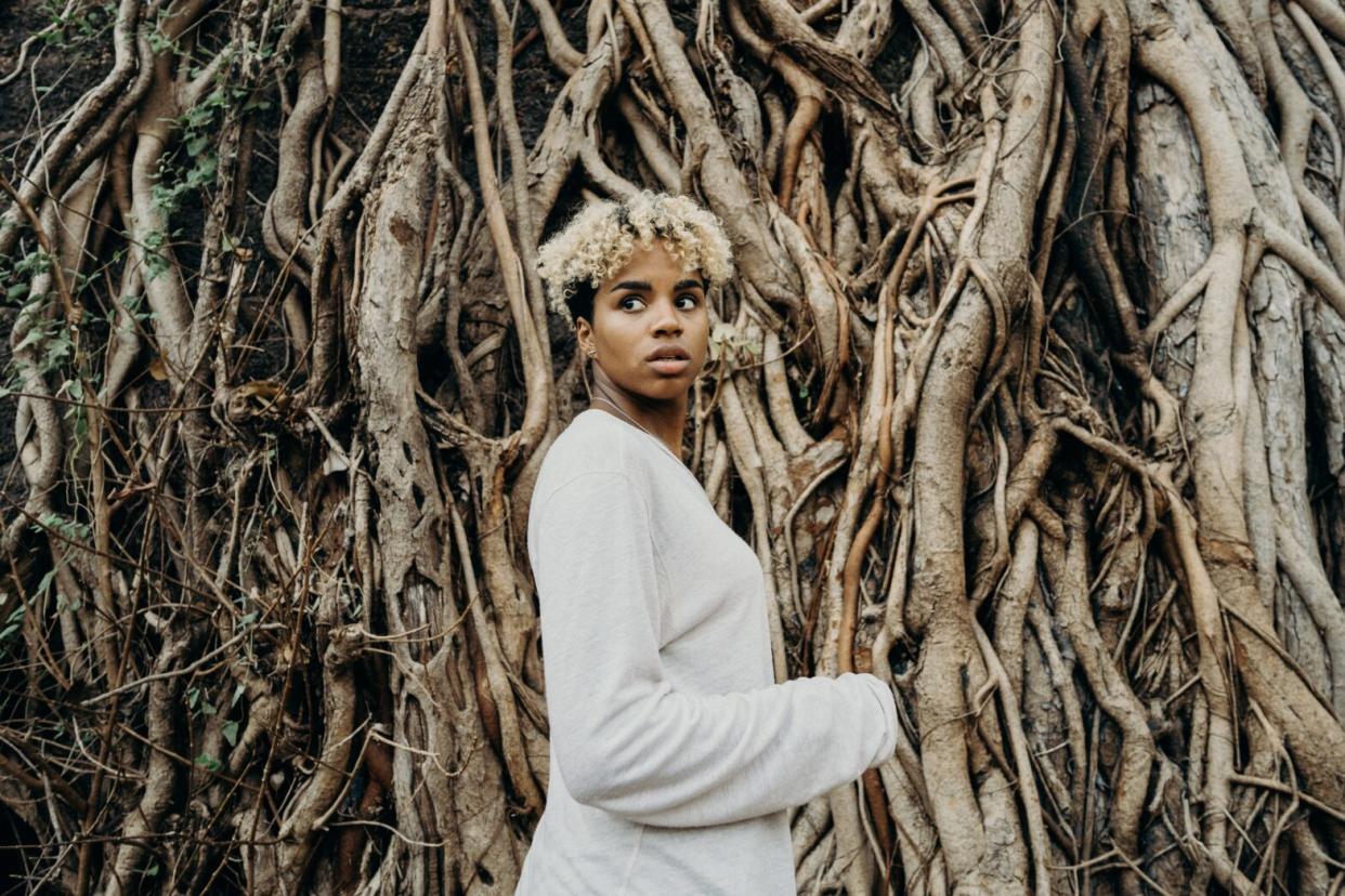 Black woman with blonde hair standing in front of trees wearing beige sweater