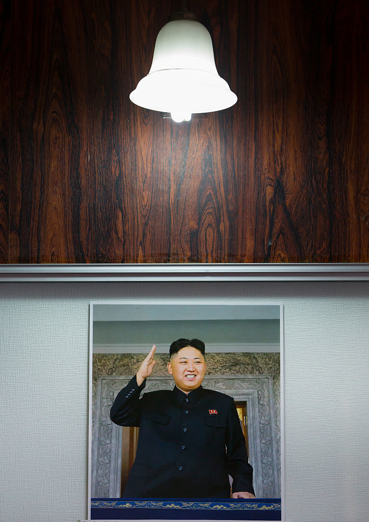 A framed photo of Kim Jong Un is displayed on a wall under a lit ceiling lamp. Kim Jong Un is pictured smiling and giving a salute