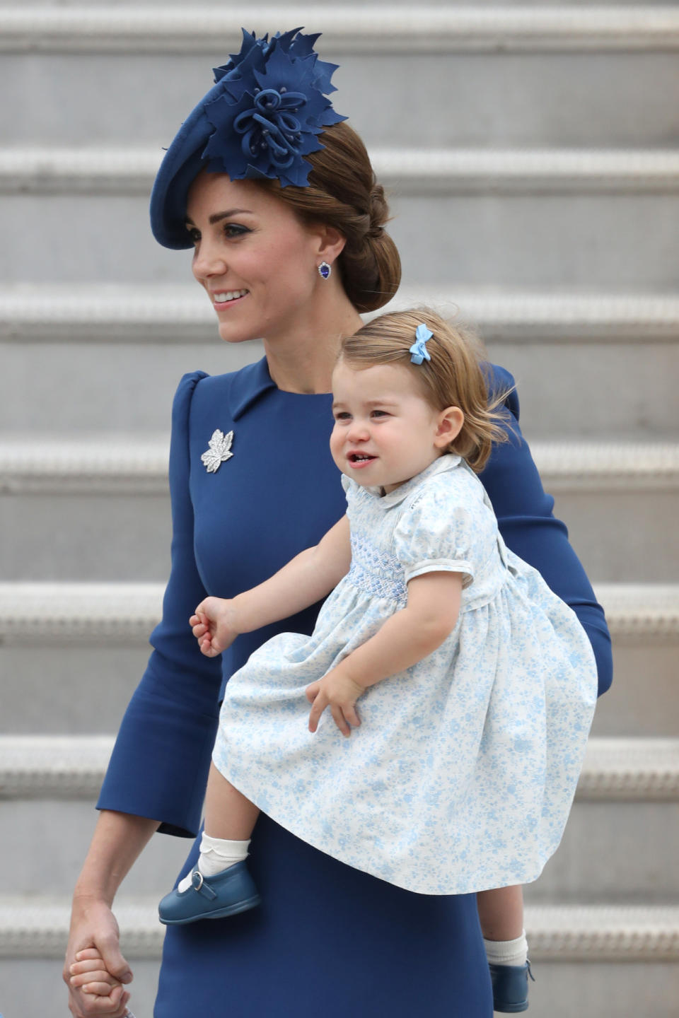 The Duchess of Cambridge in blue dress and carrying Princess Charlotte in a floral dress. 