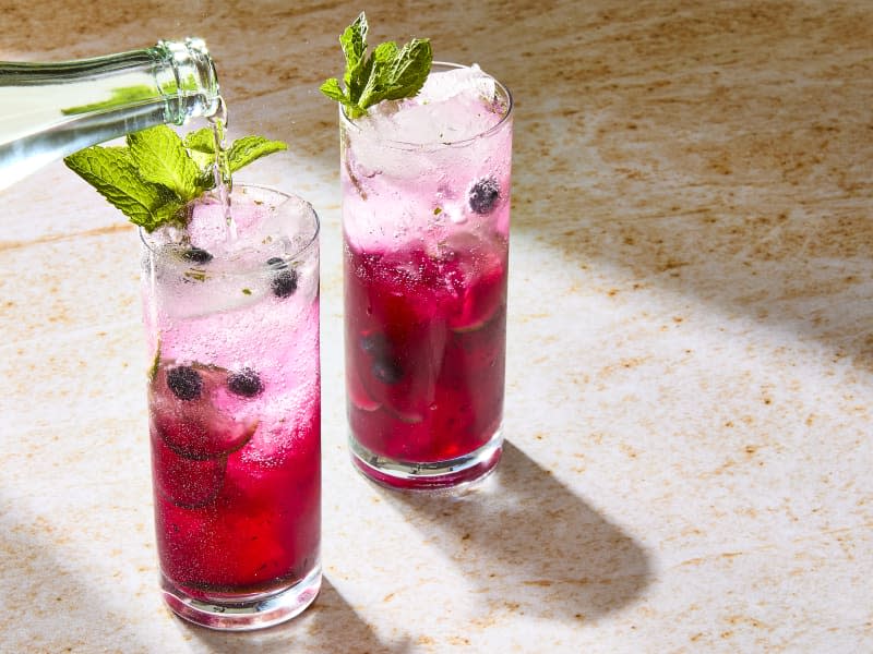 Two blueberry mojito cocktails in collins glasses with fresh blueberries and slices of lime floating in the glass, garnished with fresh mint with one being topped off with Topo Chico sparkling water.