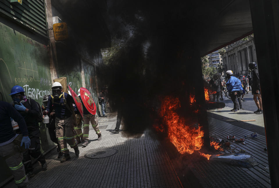 Protesters who volunteer to work as first-aid responders walk past a bus stop set fire by protesters after police blocked the route of a march against the commemoration of the discovery of the Americas that would have passed the presidential palace in Santiago, Chile, Monday, Oct. 12, 2020. The march was organized by Indigenous groups demanding autonomy and the recovery of ancestral land. (AP Photo/Esteban Felix)