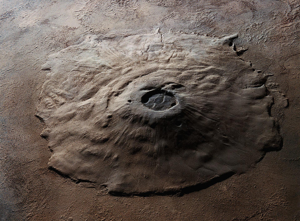 A large, circular, and raised volcanic structure with a central crater, resembling a shield volcano. The surface appears rough and textured