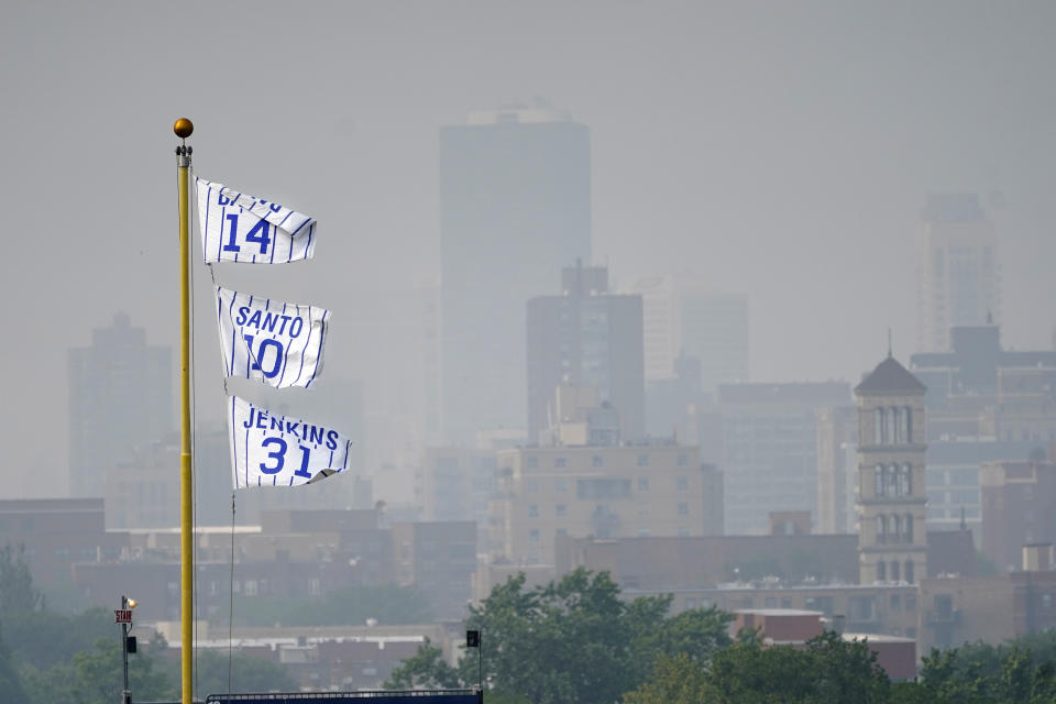 The flags of Chicago Cubs' Hall of Fame players Ernie Banks (14) Ron Santo (10) and Ferguson Jenkins fly off the Wrigley Field left field foul pole as a veil of haze from Canadian wildfires shroud high rise buildings along Lake Michigan before a baseball game between the Cubs and the Philadelphia Phillies Tuesday, June 27, 2023, in Chicago. (AP Photo/Charles Rex Arbogast)