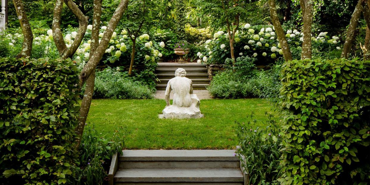 lush garden with low stairway leading to a small patch of lawn with a statue at center and trees and flowering bushes and other hedges flanking it and beyond