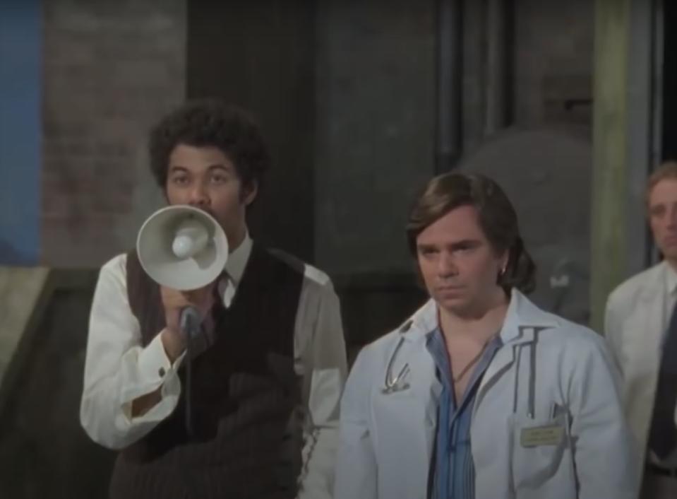 Richard Ayoade with a microphone standing on a roof next to Matt Berry on Garth Marenghi's Darkplace