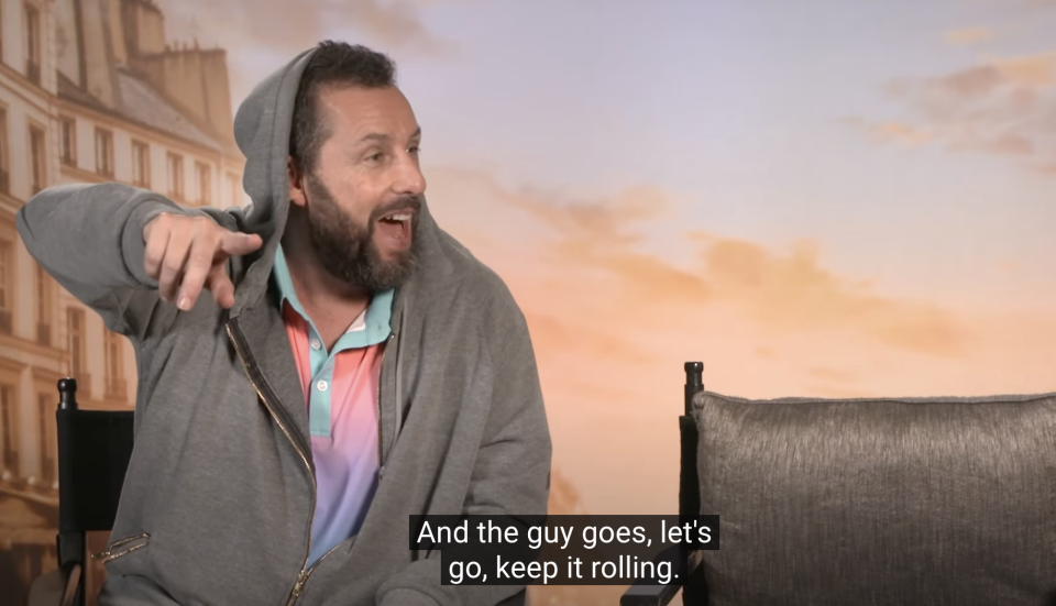 Adam Sandler, in casual attire with a hoodie, points and laughs during an interview. Caption: 