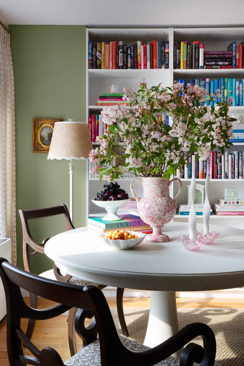 Vintage white and pink vase with large pink floral arrangement in center of table in front of color coded book shelf, round white table with vintage wood chairs