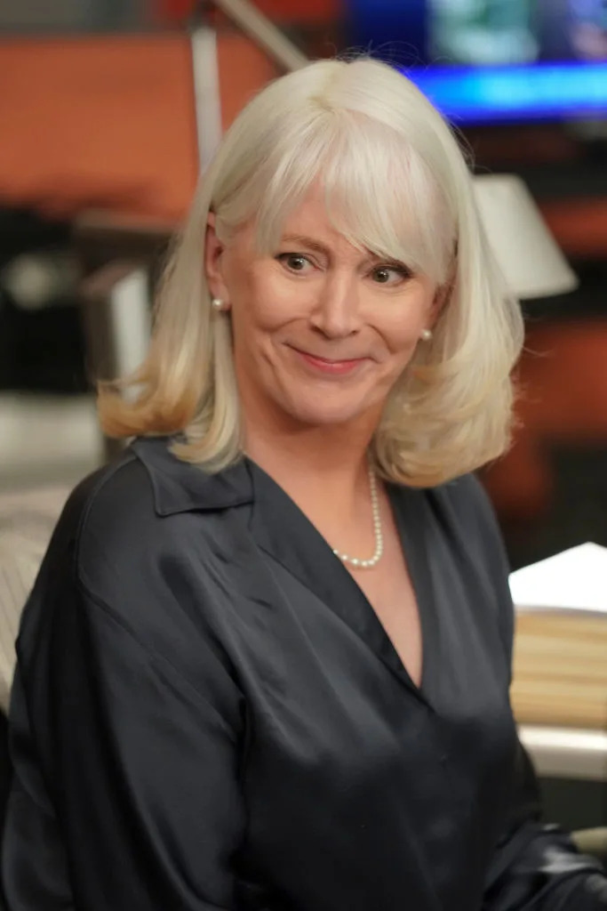 Woman in TV show scene wearing a dark blouse with pearls, sitting indoors, smiling