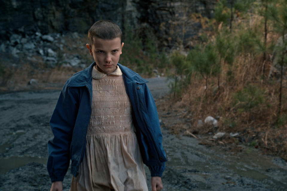 A child in a dirty, pale pink dress with a denim jacket, sporting a shaved head, nose bleed, and menacing expression; still from "Stranger Things"