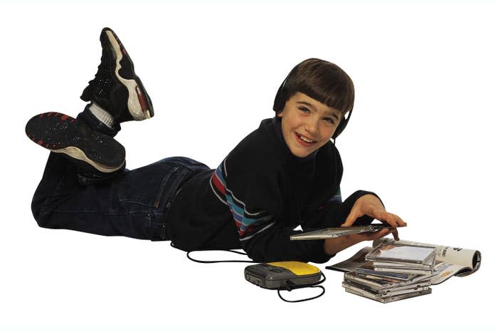 A '90s kids lying on the floor listening to a CD from his yellow portable CD player while he has a CD case opened in his hand and has a stack of CDs next to him