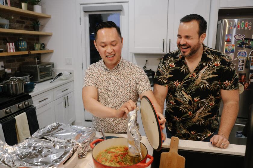 Archie Cubarrubia, left, and T.J. Morales, right, stand behind a kitchen counter. Cubarrubia lifts the lid from a red pot.