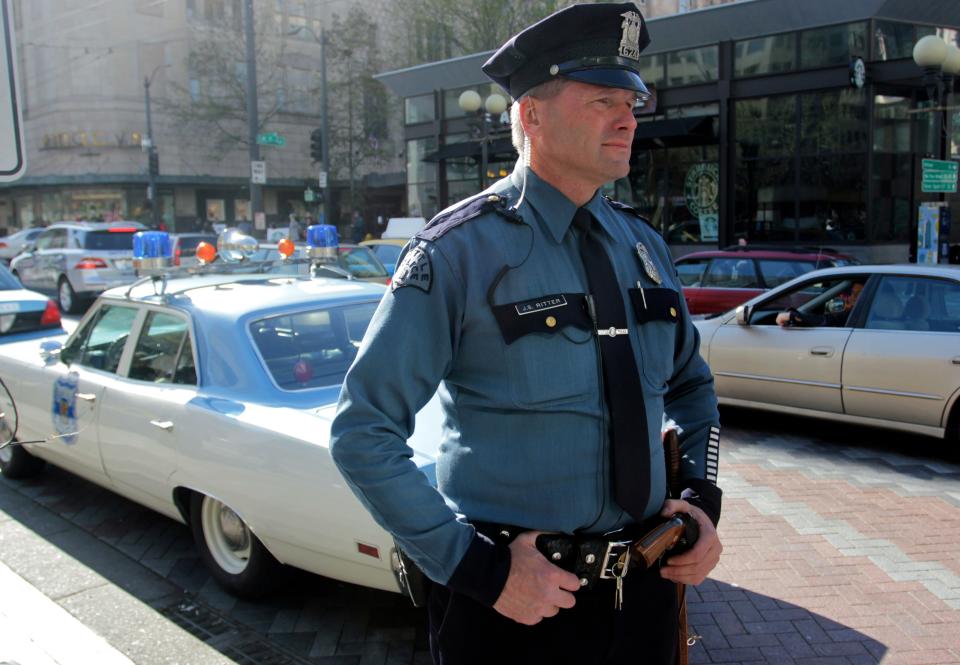 Jim Ritter stands in his police uniform in front of his 1970 Plymouth Satellite patrol car on the street.
